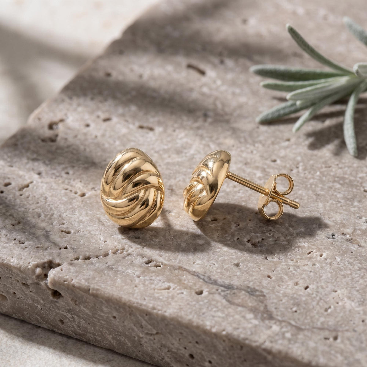 14K yellow gold braided knot stud earrings pair on a stone slab with a sprig of lavender, side view of post