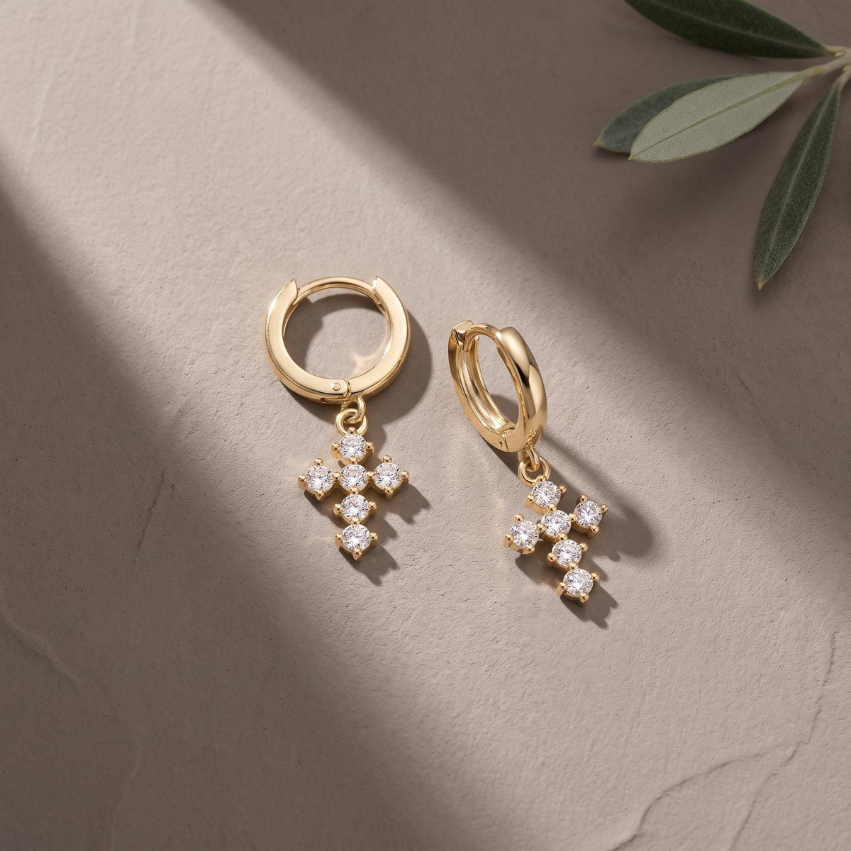 Pair of 14K yellow gold cross huggie earrings with cubic zirconia on a beige surface beside an olive leaf