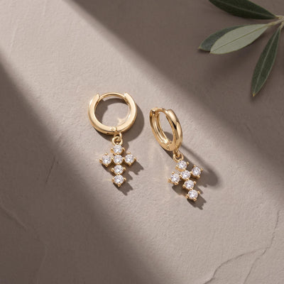 Pair of 14K yellow gold cross huggie earrings with cubic zirconia on a beige surface beside an olive leaf