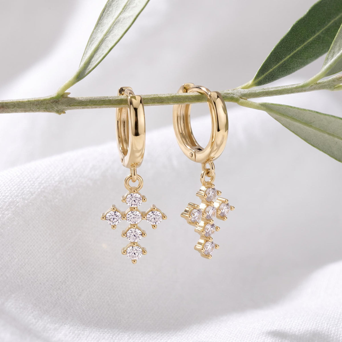 14K yellow gold cross huggie earrings with cubic zirconia hanging from an olive branch over white fabric