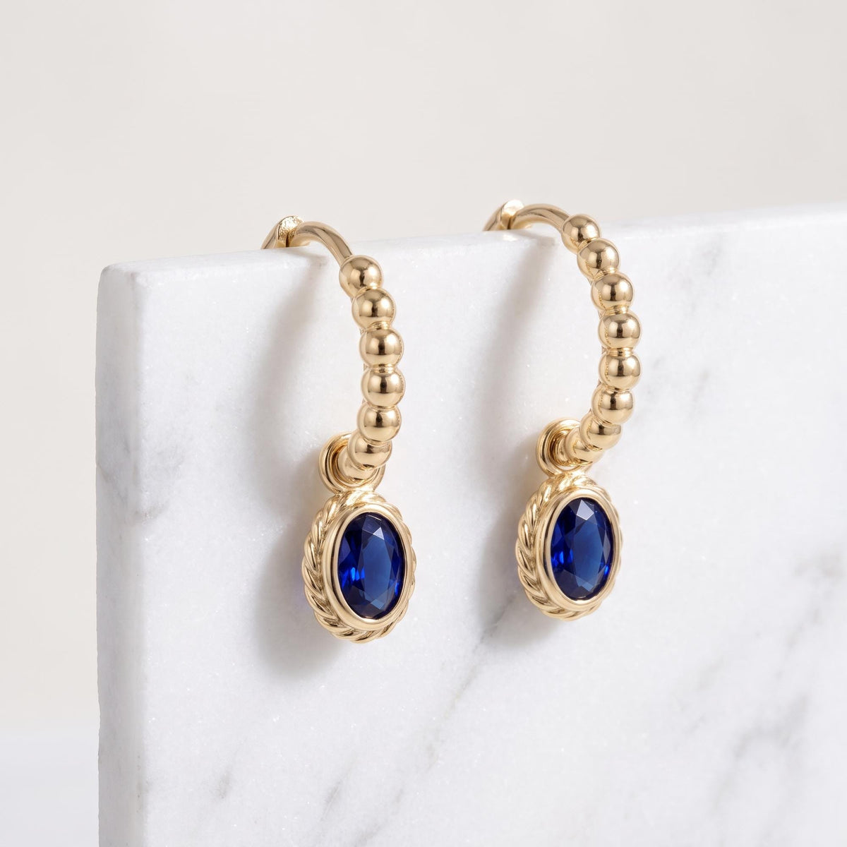 14K yellow gold beaded huggie earrings with oval blue stone drops resting on a white marble slab