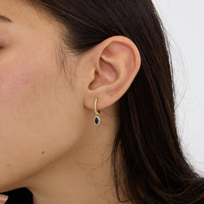 Close-up of ear wearing 14K gold beaded huggie earring with small oval blue stone drop, side profile