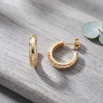 14K yellow gold cubic zirconia hoop earrings on weathered wood beside a eucalyptus leaf