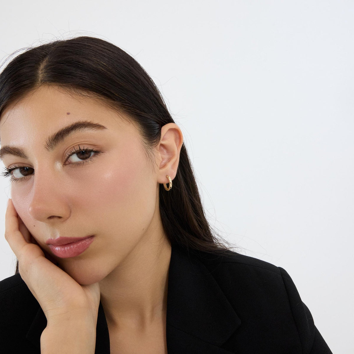 Model resting cheek on hand wearing 14K gold cubic zirconia hoop earring, black blazer, white backdrop