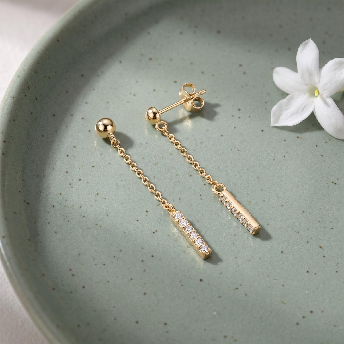 14K yellow gold drop stud earrings with cubic zirconia bars on a green speckled dish beside a white flower