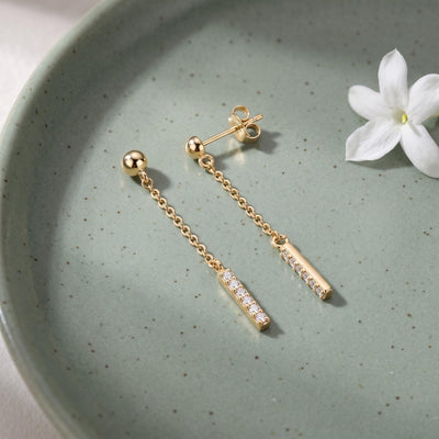 14K yellow gold drop stud earrings with cubic zirconia bars on a green speckled dish beside a white flower