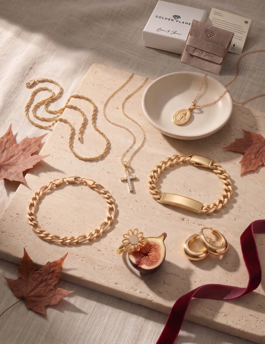 14K gold Figaro and Cuban ID bracelets, cross and medal necklaces, daisy ring and dome hoop earrings on travertine with autumn maple leaves