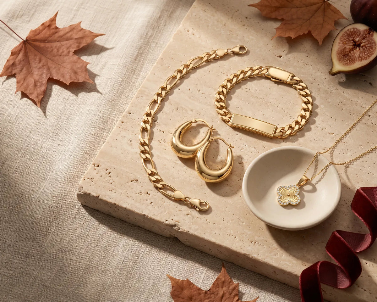 14K gold Figaro bracelet, Cuban ID bracelet, dome hoop earrings and clover necklace on travertine with autumn leaves