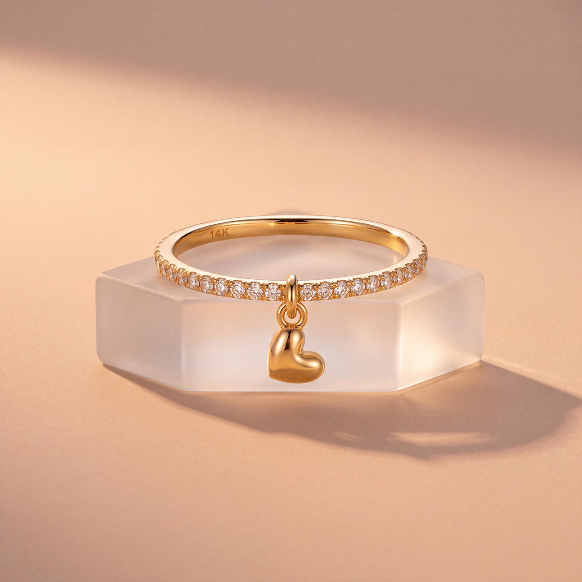 14K gold heart charm ring resting on a frosted hexagon block in warm peach light