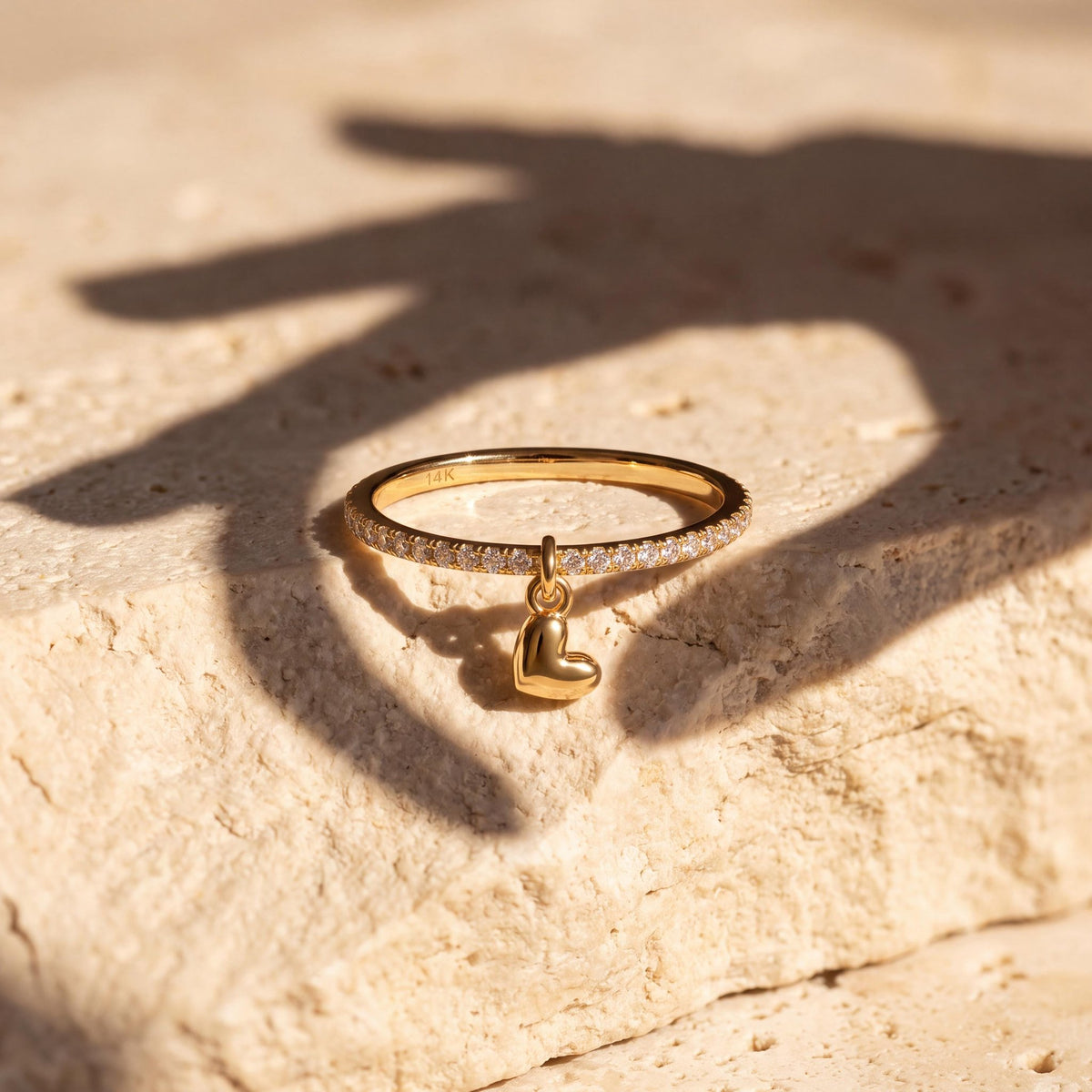 14K yellow gold heart charm ring on a limestone block with a hand's shadow falling across it