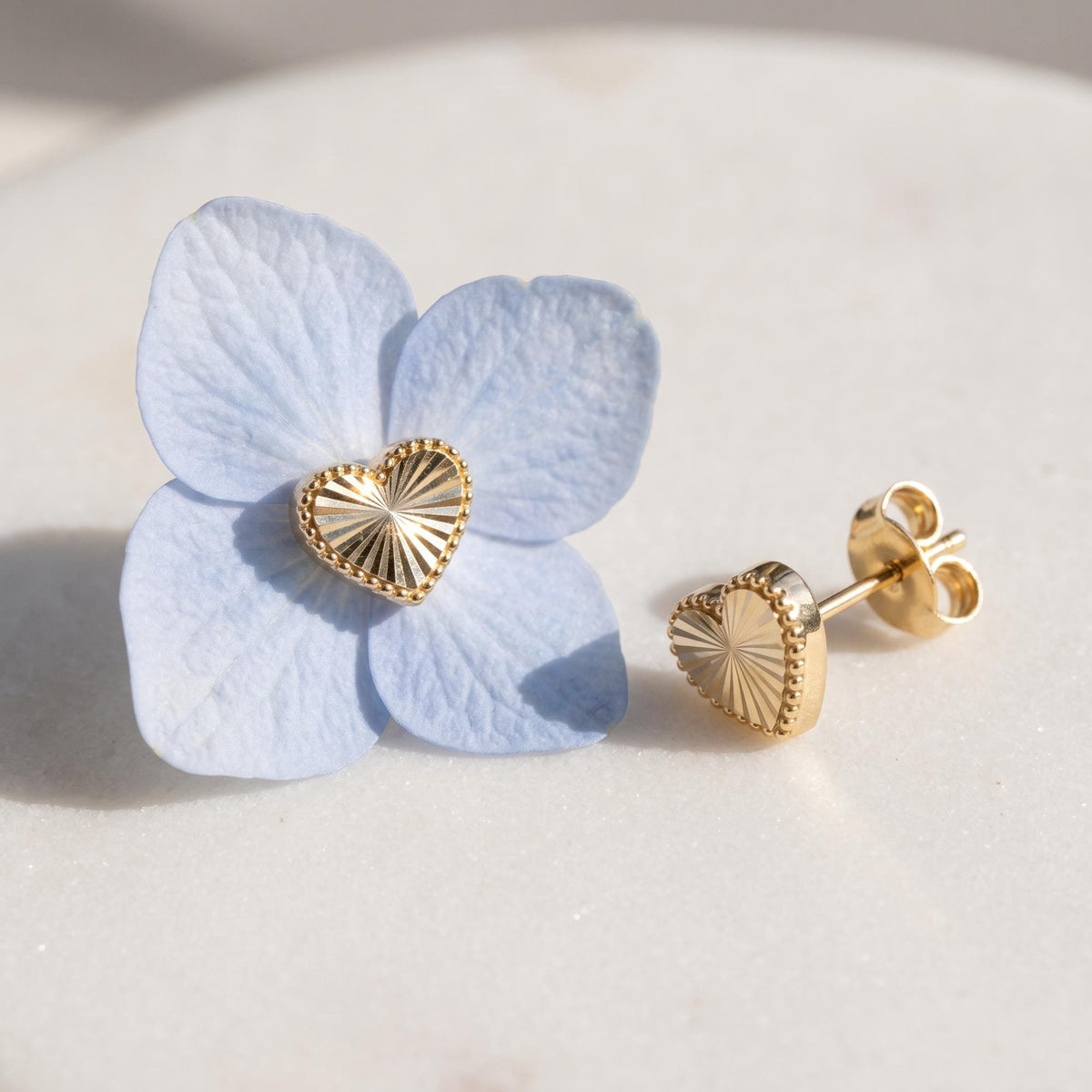 14K gold heart stud earrings with sunburst texture beside a pale blue hydrangea petal on white marble