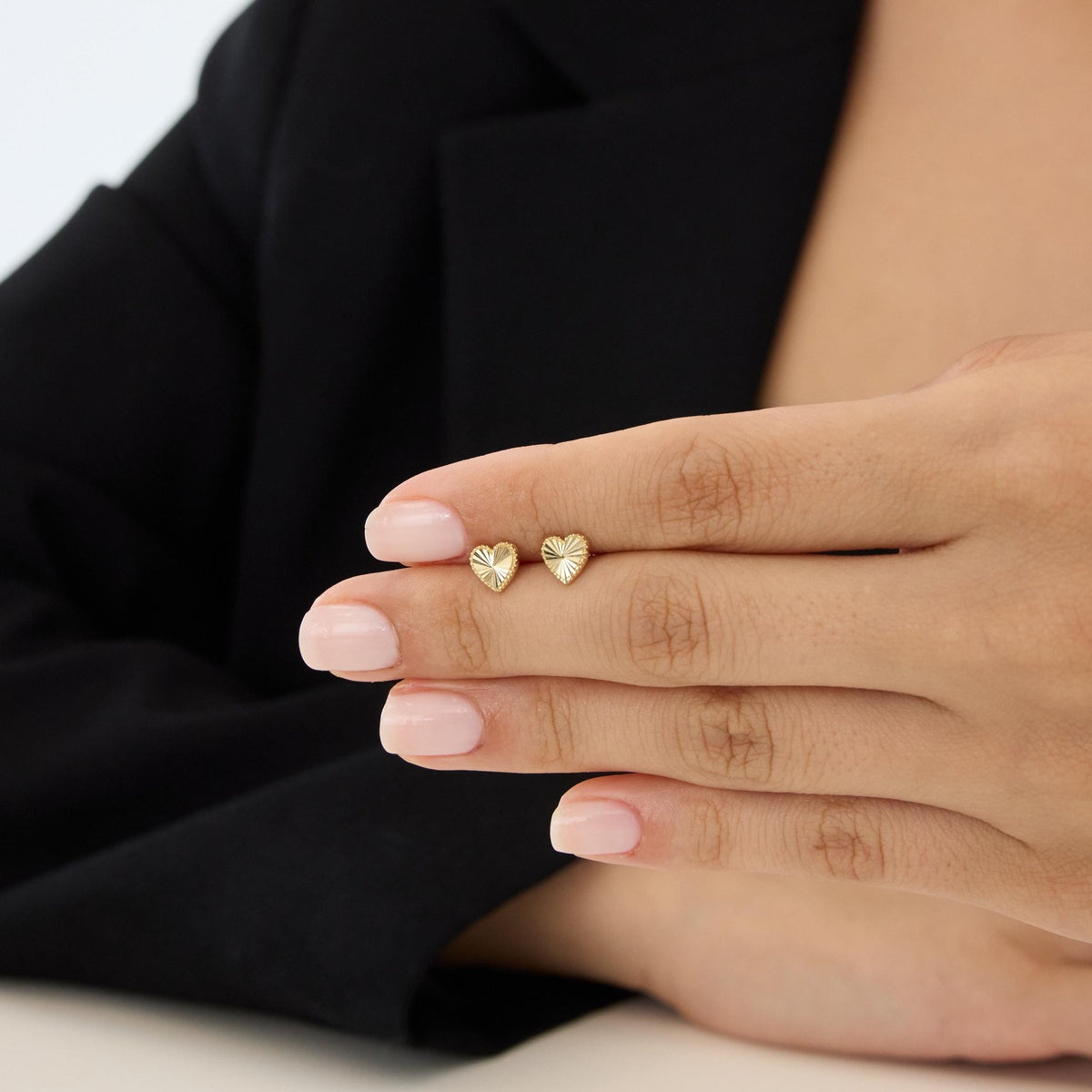 14K gold heart stud earrings with ray-cut finish held on a woman's fingertips, black blazer behind