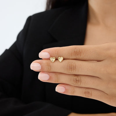 Pair of 14K gold heart stud earrings resting on manicured fingers, close view with dark blazer background