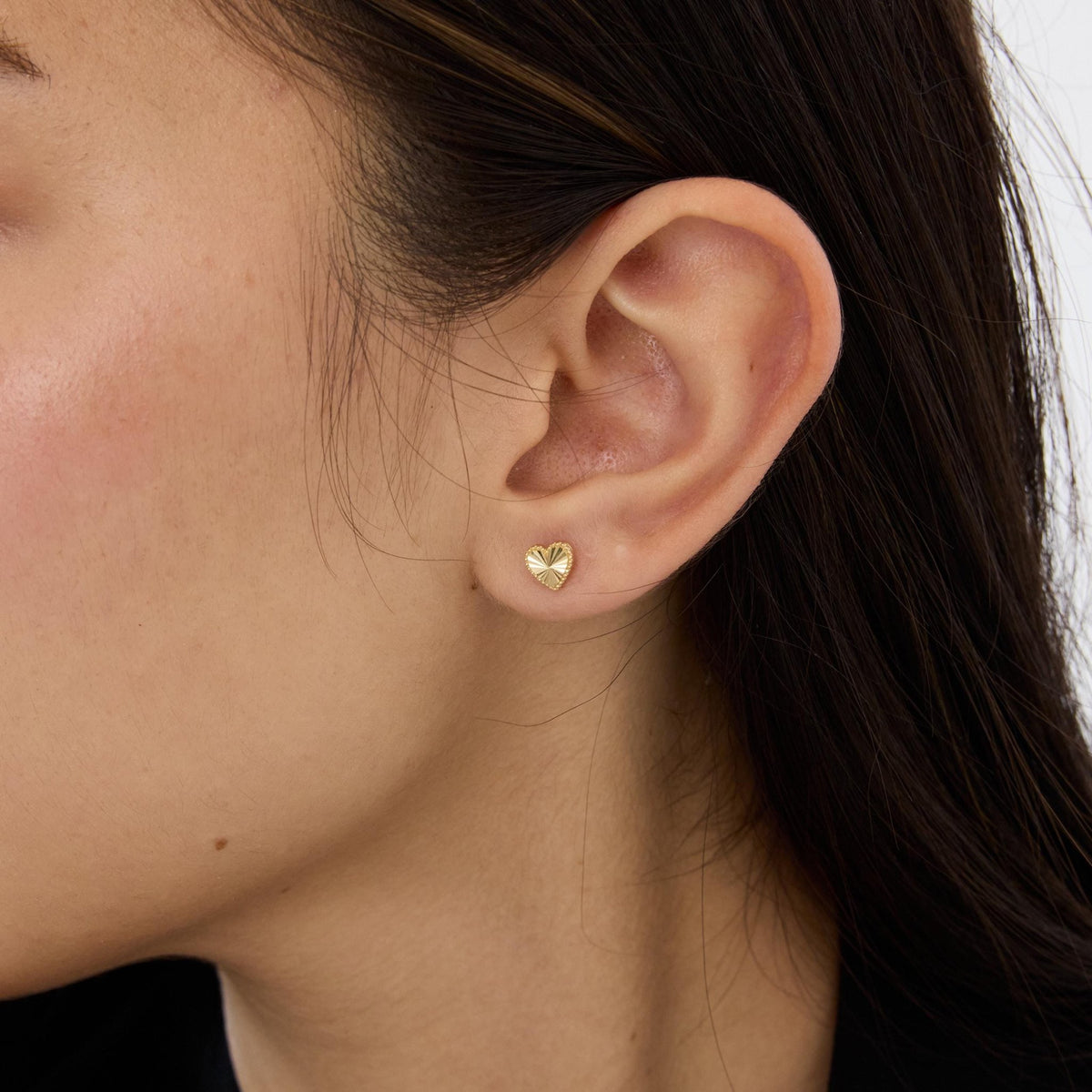 Close-up of a 14K gold heart stud earring worn on the ear, ray-cut face and dark hair in view