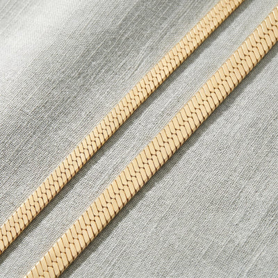 Close-up of two 14K yellow gold herringbone chains lying flat diagonally on grey linen fabric