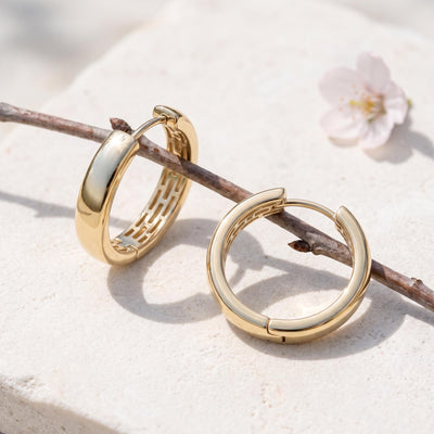 14K yellow gold huggie hoop earrings resting on a pale stone beside a bare branch and blossom