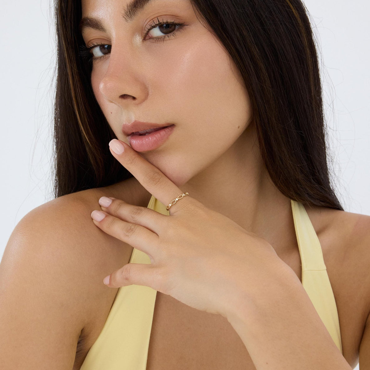 14K gold infinity ring on the index finger, model glancing sideways with fingertip at her lips