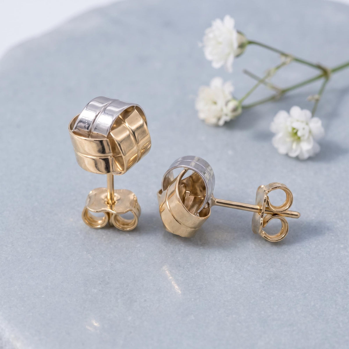 14K yellow gold knot stud earrings with white gold accent strip, resting on grey stone beside small white flowers