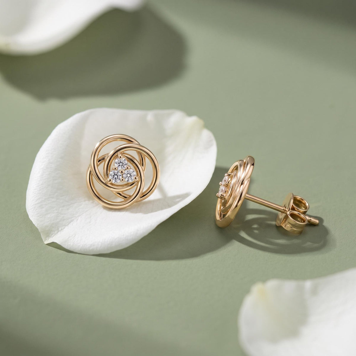 14K yellow gold love knot stud earrings with cubic zirconia, resting on a white petal, side view showing post