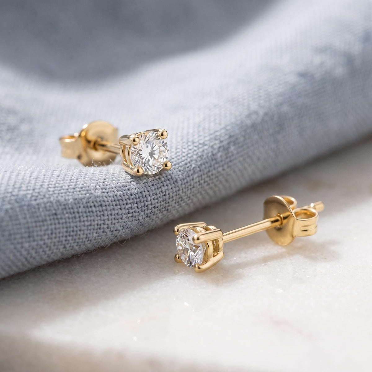 14K yellow gold round cubic zirconia solitaire stud earrings, pair resting on grey linen and white marble
