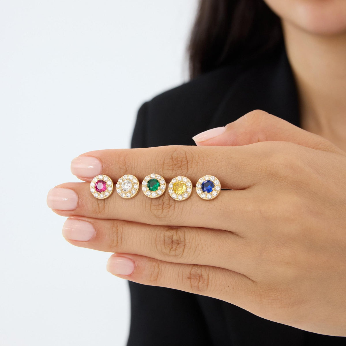 14K gold round halo stud earrings in five stone colors lined up across a model's fingers