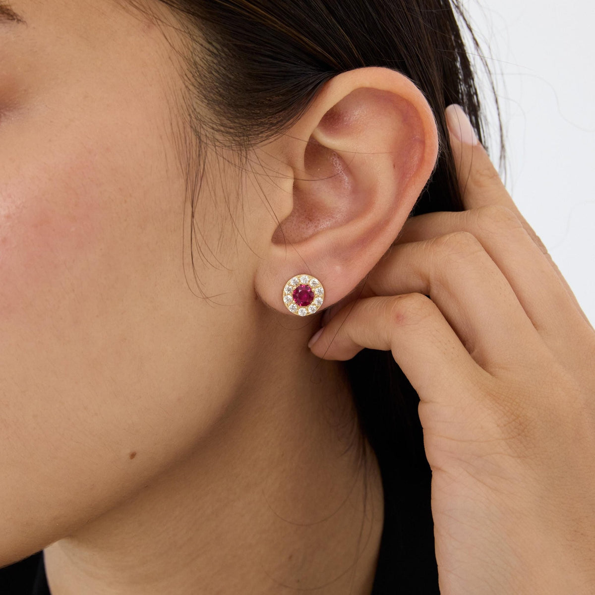 Close-up of 14K gold round halo stud earring with simulated ruby worn on a model's ear