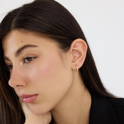 Woman resting chin on hand wearing a 14K gold square huggie hoop earring with cubic zirconia