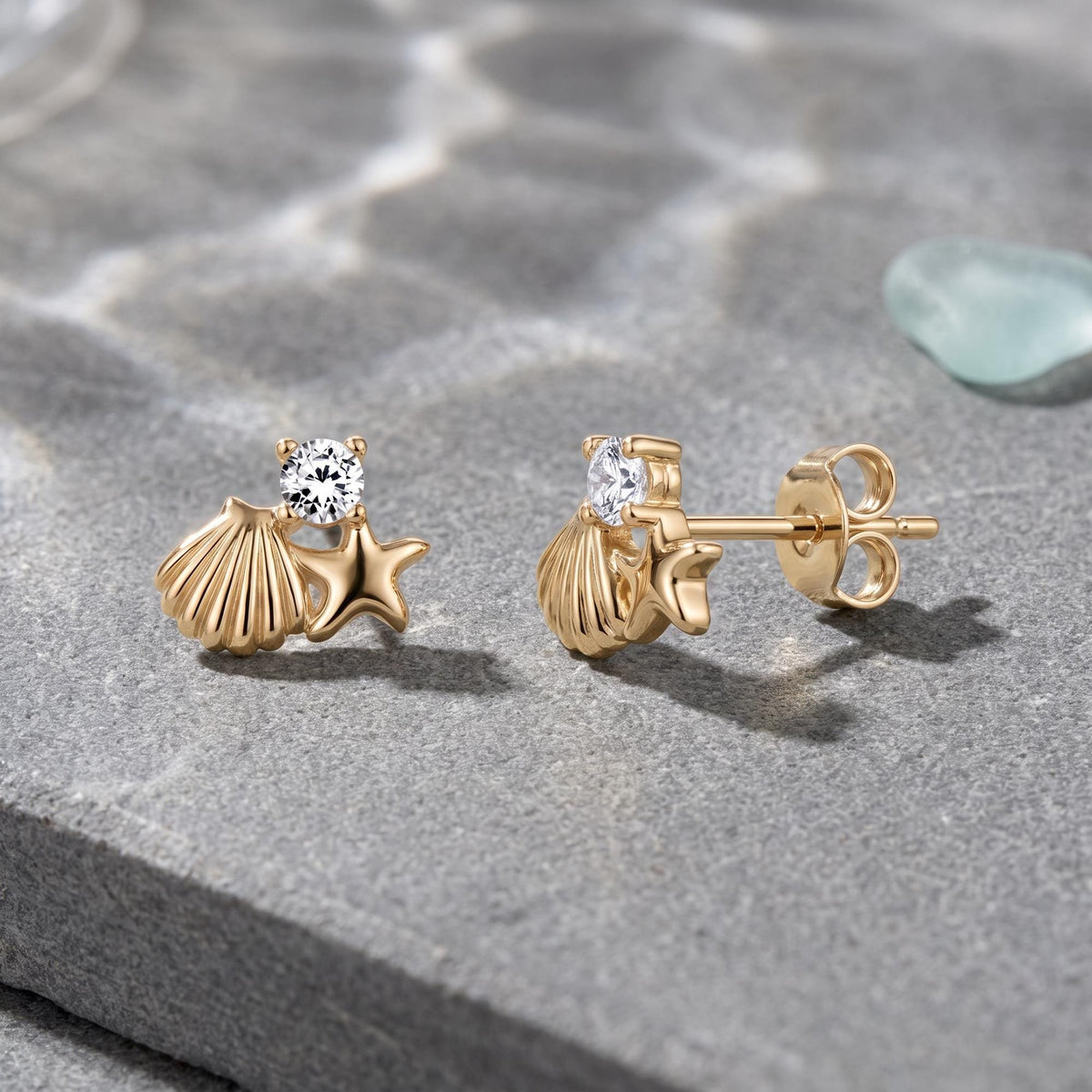 14K gold starfish and shell stud earrings with round cubic zirconia on grey stone beside sea glass