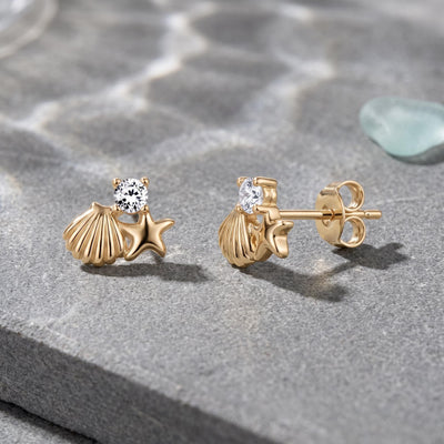 14K gold starfish and shell stud earrings with round cubic zirconia on grey stone beside sea glass