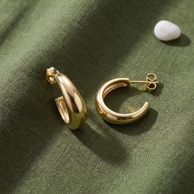 14K yellow gold tapered hoop earrings, one pair with post backs on green linen next to a white pebble