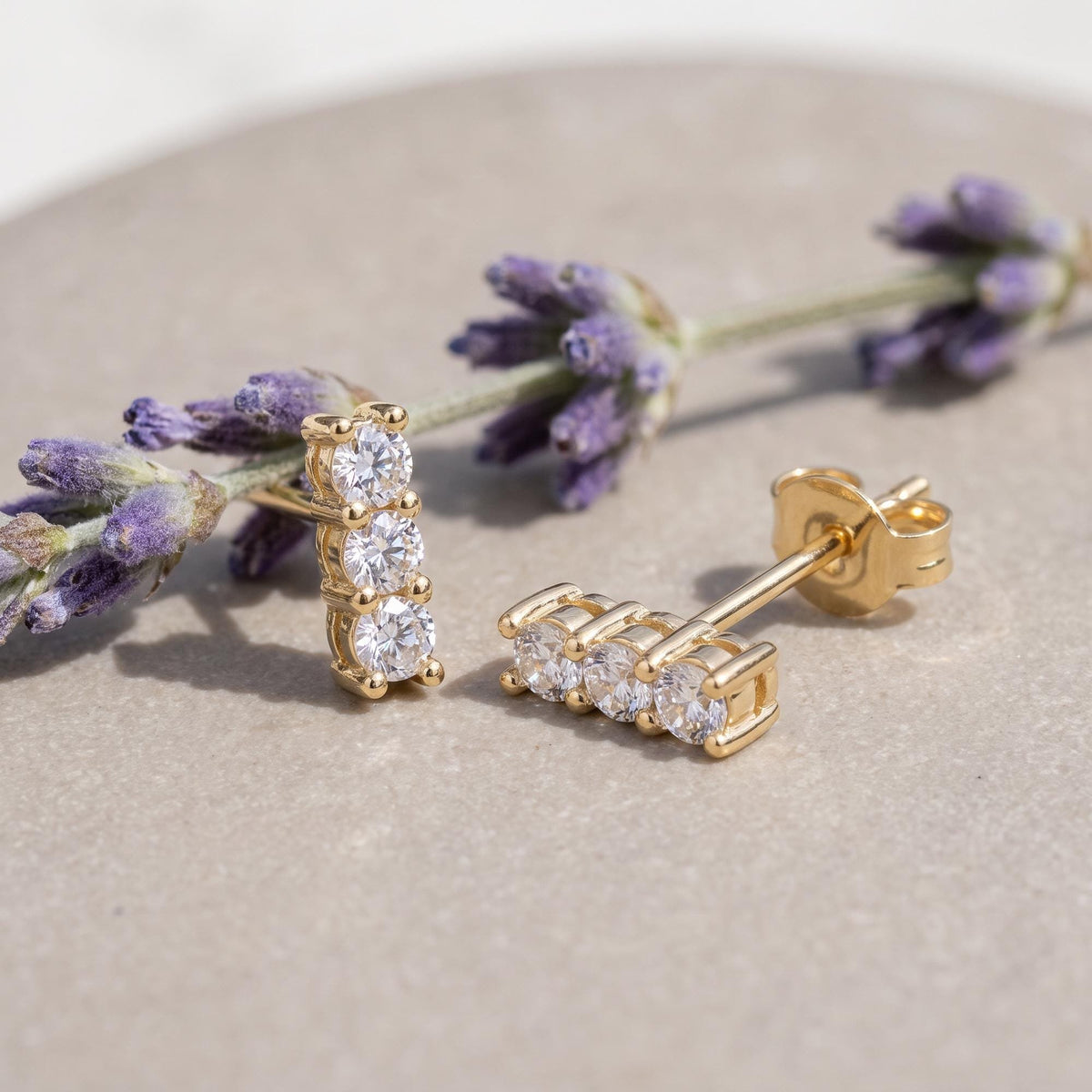 14K yellow gold three stone cubic zirconia stud earrings resting on a stone beside lavender sprigs