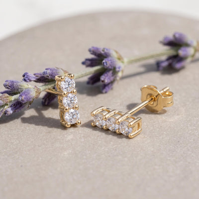 14K yellow gold three stone cubic zirconia stud earrings resting on a stone beside lavender sprigs