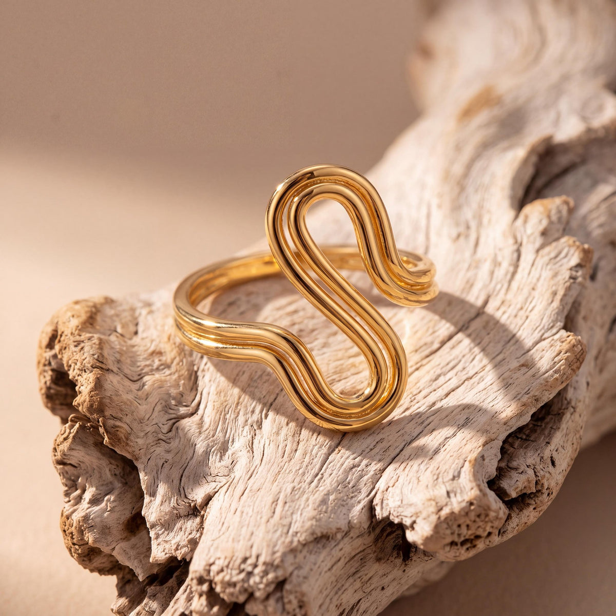 14K yellow gold wave ring standing on a piece of pale driftwood against a beige backdrop
