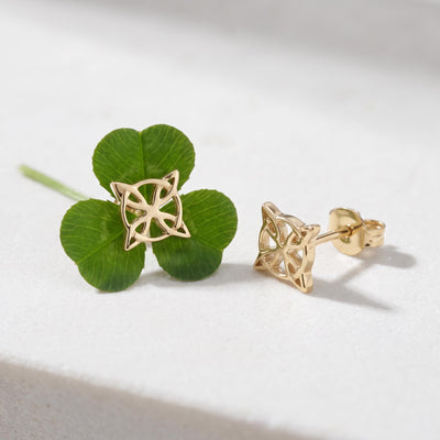 14K gold witch knot stud earrings, one resting on a four-leaf clover and one beside it on a white surface